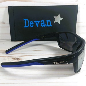 Peut inclure: Lunettes de soleil noires avec des d&eacute;tails bleus sur les branches et des verres fonc&eacute;s. Elles sont pos&eacute;es &agrave; c&ocirc;t&eacute; d'un &eacute;tui noir avec le nom "Devan" en bleu et une &eacute;toile argent&eacute;e.
