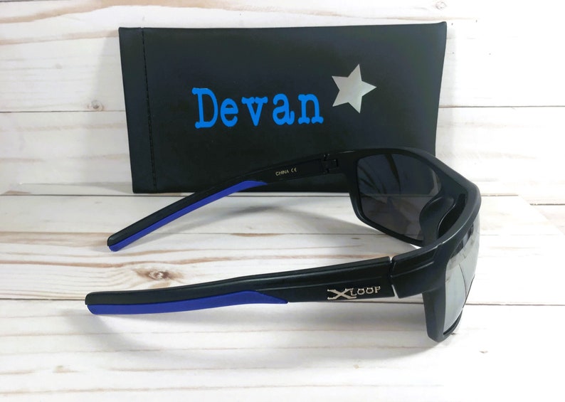 Peut inclure: Lunettes de soleil noires avec des d&eacute;tails bleus sur les branches et des verres fonc&eacute;s. Elles sont pos&eacute;es &agrave; c&ocirc;t&eacute; d'un &eacute;tui noir avec le nom "Devan" en bleu et une &eacute;toile argent&eacute;e.