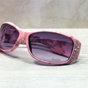 May include: Pink sunglasses with a marble pattern and rhinestones on the side. The sunglasses have a purple lens and the word "Sadie" is printed on the frame.