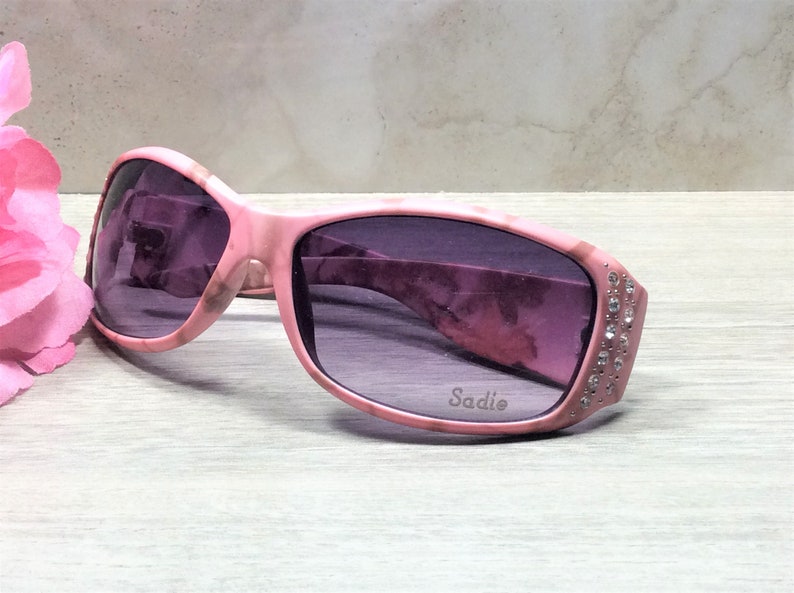 May include: Pink sunglasses with a marble pattern and rhinestones on the side. The sunglasses have a purple lens and the word "Sadie" is printed on the frame.