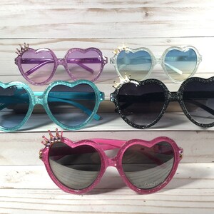 May include: Five pairs of heart-shaped sunglasses with glitter and a crown detail on the top of the frames. The sunglasses are in various colors, including pink, blue, black, and purple.