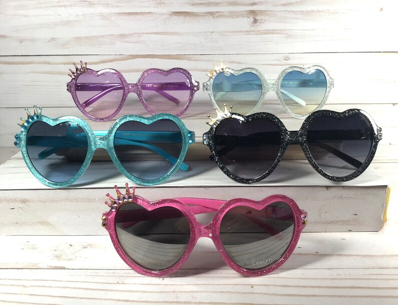 May include: Five pairs of heart-shaped sunglasses with glitter and a crown detail on the top of the frames. The sunglasses are in various colors, including pink, blue, black, and purple.