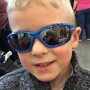 May include: A young boy wearing blue and black sunglasses with the word "Bentley" on the right lens. The sunglasses have a reflective blue lens.