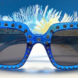 May include: Blue square sunglasses with silver studs around the frame. The sunglasses have dark lenses.