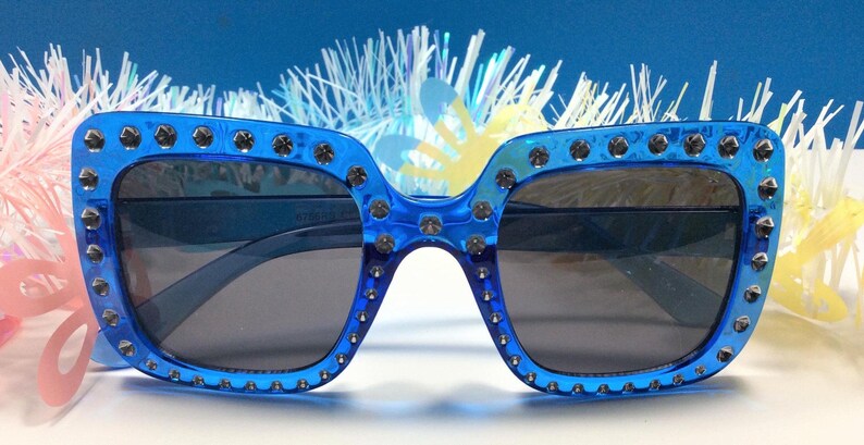 May include: Blue square sunglasses with silver studs around the frame. The sunglasses have dark lenses.