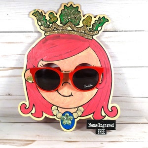 May include: A wooden cutout of a girl's face with pink hair, wearing red sunglasses with the word "Camilla" on the right lens. The girl is wearing a gold necklace with a green four-leaf clover pendant. The cutout is decorated with a gold crown with green leaves and the text "Name Engraved FREE".