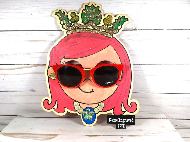 May include: A wooden cutout of a girl's face with pink hair, wearing red sunglasses with the word "Camilla" on the right lens. The girl is wearing a gold necklace with a green four-leaf clover pendant. The cutout is decorated with a gold crown with green leaves and the text "Name Engraved FREE".