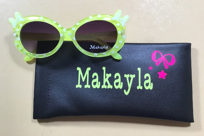 May include: Green polka dot sunglasses with a white bow on each side. The sunglasses come with a black case with the name Makayla written in green and a pink bow and star.