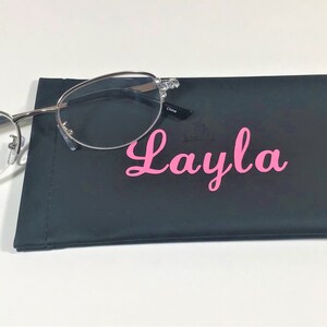 K&ouml;nnte beinhalten: Silberfarbene Brille mit klaren Gl&auml;sern liegt auf einem schwarzen Brillenetui. Das Etui ist mit dem Namen "Layla" in rosa Schrift personalisiert. Die Brille hat eine Vergr&ouml;&szlig;erung von +1,50.