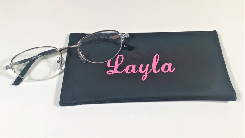K&ouml;nnte beinhalten: Silberfarbene Brille mit klaren Gl&auml;sern liegt auf einem schwarzen Brillenetui. Das Etui ist mit dem Namen "Layla" in rosa Schrift personalisiert. Die Brille hat eine Vergr&ouml;&szlig;erung von +1,50.