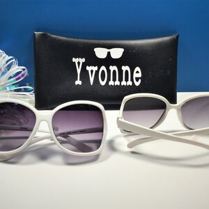May include: Two pairs of white-framed sunglasses with gradient lenses are displayed. A black case with the name "Yvonne" in white lettering and a decorative bow are in the background. The sunglasses are a classic, oversized style.