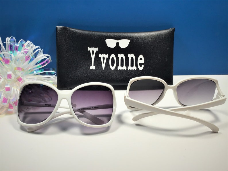 May include: Two pairs of white-framed sunglasses with gradient lenses are displayed. A black case with the name "Yvonne" in white lettering and a decorative bow are in the background. The sunglasses are a classic, oversized style.