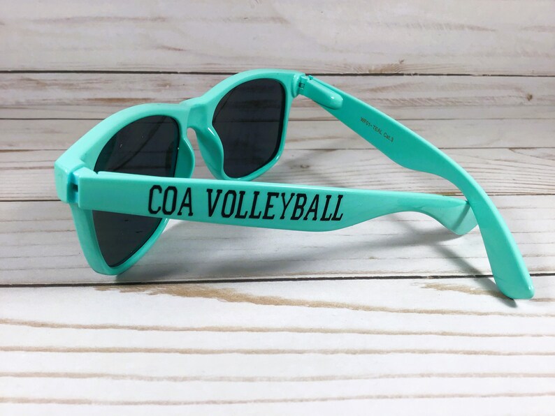 Puede incluir: Gafas de sol de color verde azulado con lentes oscuras que presentan el texto negro COA VOLLEYBALL impreso en la patilla exterior. La patilla interior incluye el texto WP01-TEAL GALS. La montura tiene una forma cl&aacute;sica con acabado mate.