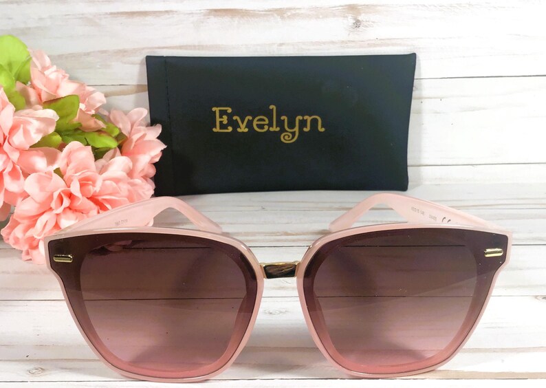 K&ouml;nnte beinhalten: Rosa Sonnenbrille mit goldenem Rahmen und braunen Gl&auml;sern. Die Sonnenbrille liegt auf einer Holzoberfl&auml;che mit einer rosa Blume im Hintergrund. Die Sonnenbrille wird mit einem schwarzen Etui geliefert, auf dem "Evelyn" in goldenen Buchstaben steht.