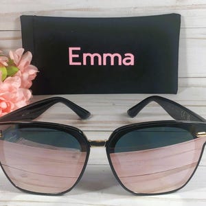 May include: Black sunglasses with rose gold mirrored lenses. The sunglasses are in a black case with the word "Emma" printed on it.