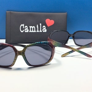 May include: Two pairs of sunglasses, one with a black frame and the other with a colorful zebra print frame. The sunglasses are on a white surface with a black case that says "Camila" with a red heart.