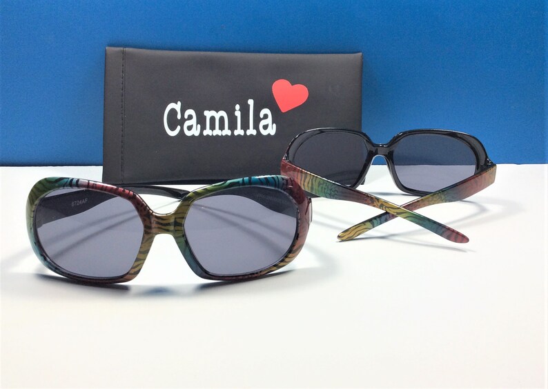 May include: Two pairs of sunglasses, one with a black frame and the other with a colorful zebra print frame. The sunglasses are on a white surface with a black case that says "Camila" with a red heart.