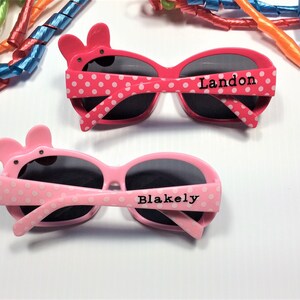 Puede incluir: Dos pares de gafas de sol rosas con lunares blancos. Las gafas tienen un bonito dise&ntilde;o de animal de dibujos animados en el lateral. Un par dice "Landon" y el otro dice "Blakely".