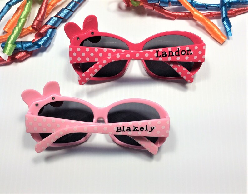 Puede incluir: Dos pares de gafas de sol rosas con lunares blancos. Las gafas tienen un bonito dise&ntilde;o de animal de dibujos animados en el lateral. Un par dice "Landon" y el otro dice "Blakely".