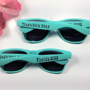 Peut inclure: Deux paires de lunettes de soleil bleu turquoise avec des verres noirs. Les lunettes de soleil ont le texte "Tina's 50th Bday" et "Florida 2022" imprim&eacute; sur les branches.