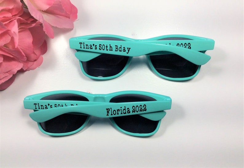 Peut inclure: Deux paires de lunettes de soleil bleu turquoise avec des verres noirs. Les lunettes de soleil ont le texte "Tina's 50th Bday" et "Florida 2022" imprim&eacute; sur les branches.