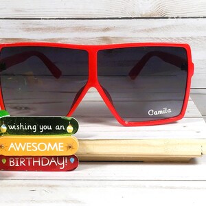 K&ouml;nnte beinhalten: Rote quadratische Sonnenbrille mit dunklem Glas. Die Sonnenbrille tr&auml;gt den Namen "Camila" auf dem rechten Glas. Die Sonnenbrille liegt auf einer Holzoberfl&auml;che mit einer bunten Geburtstagskarte, auf der "Wishing you an Awesome Birthday!" steht.