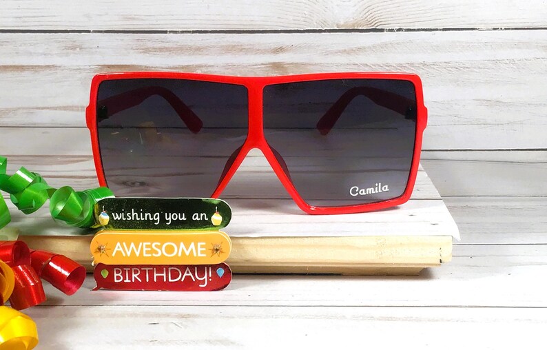 K&ouml;nnte beinhalten: Rote quadratische Sonnenbrille mit dunklem Glas. Die Sonnenbrille tr&auml;gt den Namen "Camila" auf dem rechten Glas. Die Sonnenbrille liegt auf einer Holzoberfl&auml;che mit einer bunten Geburtstagskarte, auf der "Wishing you an Awesome Birthday!" steht.