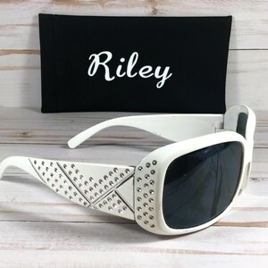 K&ouml;nnte beinhalten: Wei&szlig;e Sonnenbrille mit Strasssteinen auf dem Rahmen und dunklen Gl&auml;sern. Die Sonnenbrille liegt auf einer wei&szlig;en Holzoberfl&auml;che. Die Sonnenbrille wird mit einem schwarzen Etui geliefert, auf dem der Name "Riley" gestickt ist.