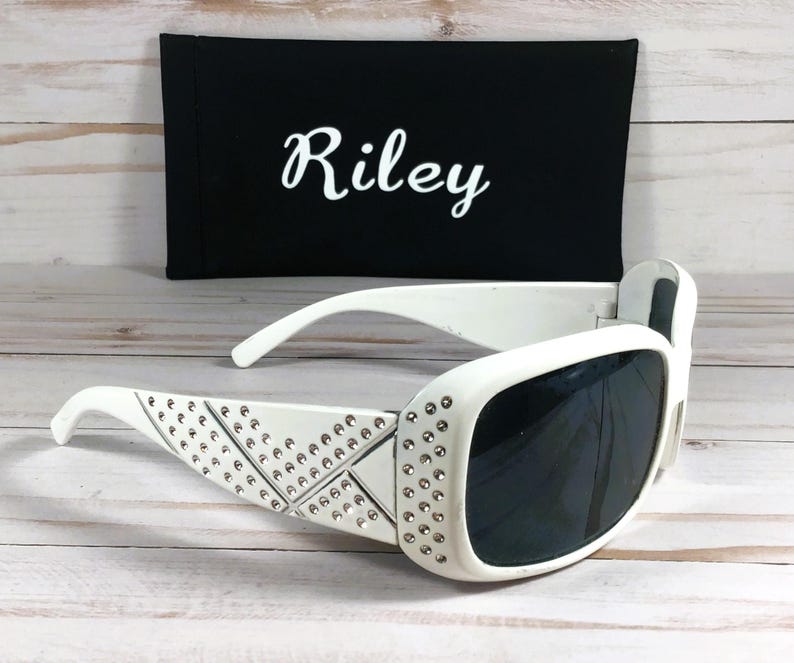 K&ouml;nnte beinhalten: Wei&szlig;e Sonnenbrille mit Strasssteinen auf dem Rahmen und dunklen Gl&auml;sern. Die Sonnenbrille liegt auf einer wei&szlig;en Holzoberfl&auml;che. Die Sonnenbrille wird mit einem schwarzen Etui geliefert, auf dem der Name "Riley" gestickt ist.