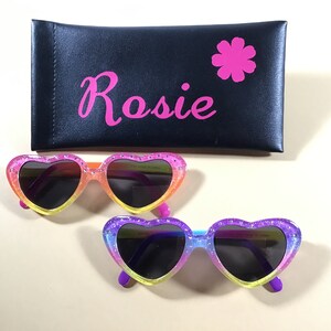 May include: A pair of heart-shaped sunglasses with a rainbow glitter design. The sunglasses have black lenses and are in a pink case with the name "Rosie" and a pink flower.