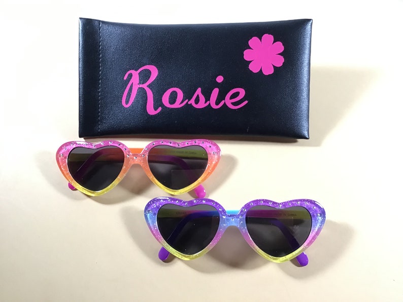 May include: A pair of heart-shaped sunglasses with a rainbow glitter design. The sunglasses have black lenses and are in a pink case with the name "Rosie" and a pink flower.