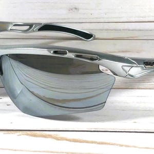May include: Silver-toned athletic sunglasses featuring a semi-rimless frame design with mirrored lenses. The temples include black rubberized grip inserts and a metallic logo plate on the side, with ventilated cutouts along the upper frame.