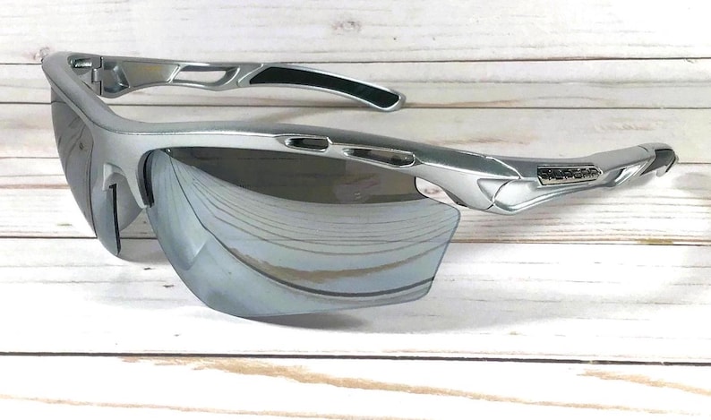 May include: Silver-toned athletic sunglasses featuring a semi-rimless frame design with mirrored lenses. The temples include black rubberized grip inserts and a metallic logo plate on the side, with ventilated cutouts along the upper frame.