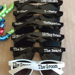 May include: A stack of black and white sunglasses with various personalized text on the frames, including "Crzy Uncl Dave", "T-Nasty", "ThePlayerTwo", "Lil Chzy Potato", "The Beard", and "The Groom".