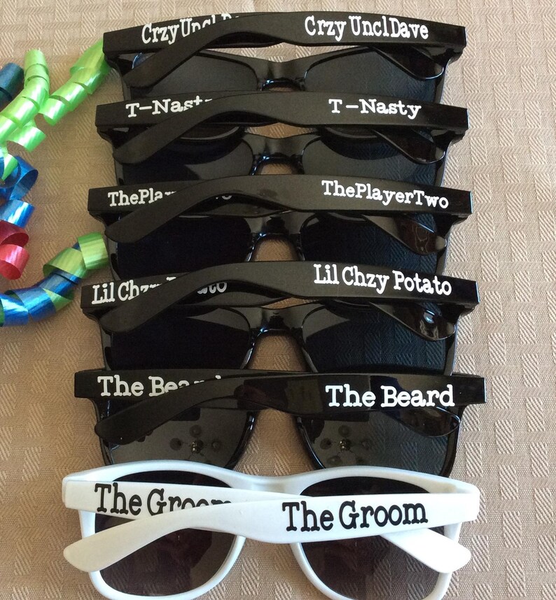 May include: A stack of black and white sunglasses with various personalized text on the frames, including "Crzy Uncl Dave", "T-Nasty", "ThePlayerTwo", "Lil Chzy Potato", "The Beard", and "The Groom".