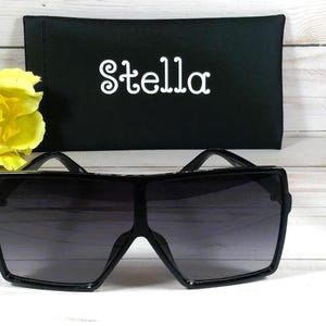 K&ouml;nnte beinhalten: Schwarze quadratische Sonnenbrille mit einem schwarzen Etui, auf dem vorne "Stella" steht.