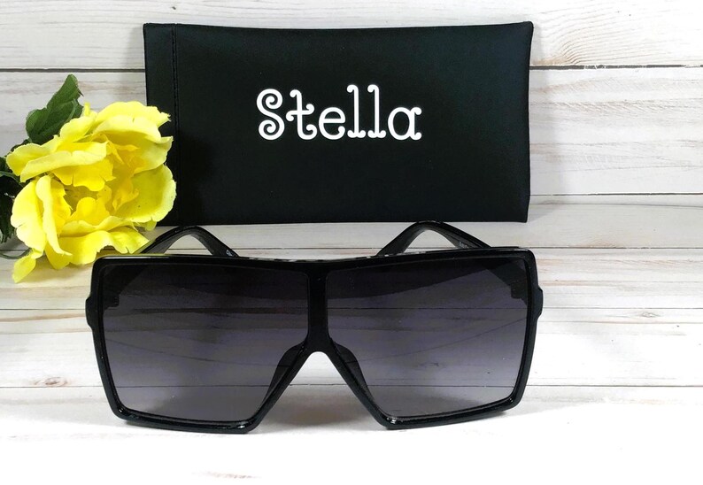 K&ouml;nnte beinhalten: Schwarze quadratische Sonnenbrille mit einem schwarzen Etui, auf dem vorne "Stella" steht.