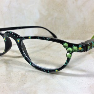 K&ouml;nnte beinhalten: Schwarze Brille mit einem floralen Muster in Gr&uuml;n, Gelb und T&uuml;rkis. Die Fassungen sind mit gr&uuml;nen Edelsteinen an den B&uuml;geln verziert. Die Gl&auml;ser sind klar.