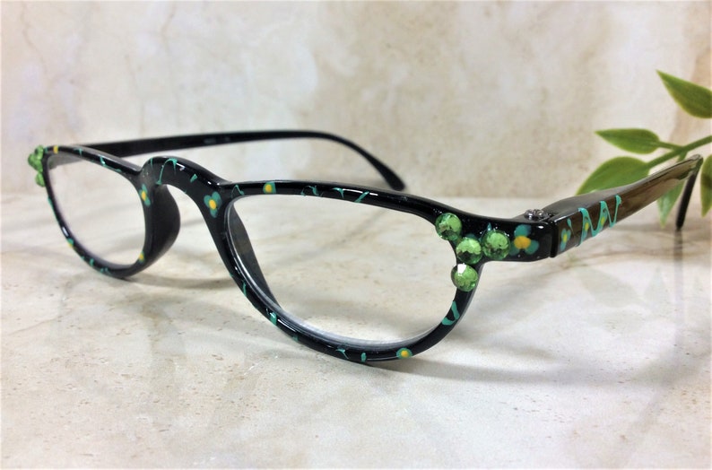 K&ouml;nnte beinhalten: Schwarze Brille mit einem floralen Muster in Gr&uuml;n, Gelb und T&uuml;rkis. Die Fassungen sind mit gr&uuml;nen Edelsteinen an den B&uuml;geln verziert. Die Gl&auml;ser sind klar.