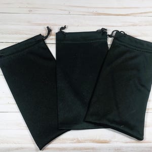 May include: Three black drawstring pouches are arranged on a light wood surface. Each pouch is rectangular with a rounded bottom and a drawstring closure at the top. The pouches are made of a soft, dark fabric.