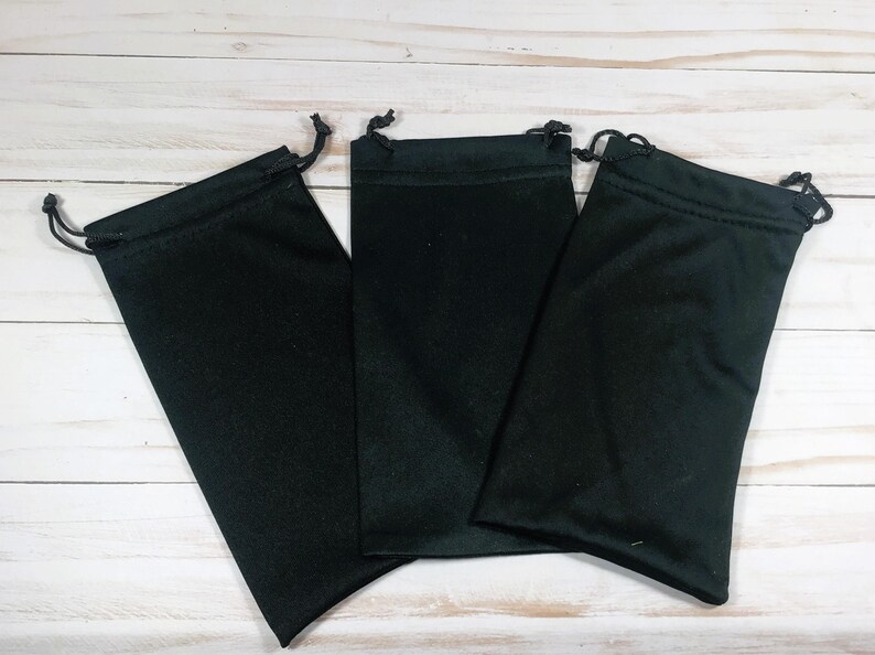 May include: Three black drawstring pouches are arranged on a light wood surface. Each pouch is rectangular with a rounded bottom and a drawstring closure at the top. The pouches are made of a soft, dark fabric.
