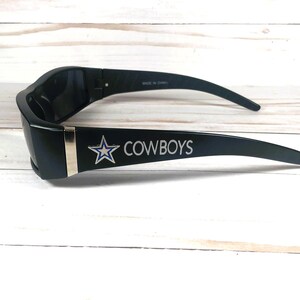 May include: Black sunglasses with the word "COWBOYS" in white lettering and a star logo. The sunglasses have a silver accent on the frame and are made in China.
