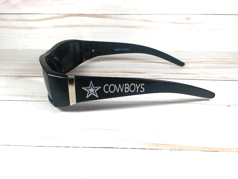 May include: Black sunglasses with the word "COWBOYS" in white lettering and a star logo. The sunglasses have a silver accent on the frame and are made in China.