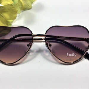 May include: Heart-shaped sunglasses with a rose gold frame and brown gradient lenses. The sunglasses have the text "UV Protection" and "Emily" printed on the lenses.