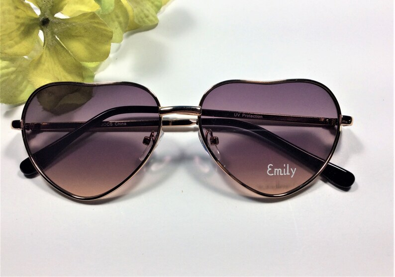 May include: Heart-shaped sunglasses with a rose gold frame and brown gradient lenses. The sunglasses have the text "UV Protection" and "Emily" printed on the lenses.