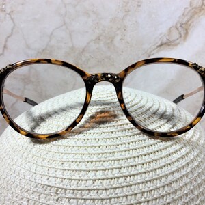 May include: Round eyeglasses with a tortoiseshell pattern frame. The frame is adorned with decorative embellishments on the outer edges. The lenses are clear, and the temples are a light brown color. The glasses are resting on a white, woven hat.