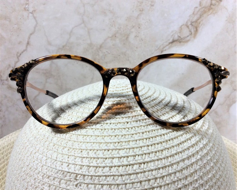 May include: Round eyeglasses with a tortoiseshell pattern frame. The frame is adorned with decorative embellishments on the outer edges. The lenses are clear, and the temples are a light brown color. The glasses are resting on a white, woven hat.