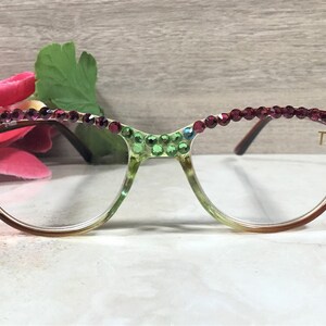 May include: Eyeglasses with a gradient frame, adorned with sparkling rhinestones in shades of green and burgundy. The lenses are clear, and the right temple features the initials "TK". The glasses are set against a soft background with floral accents.