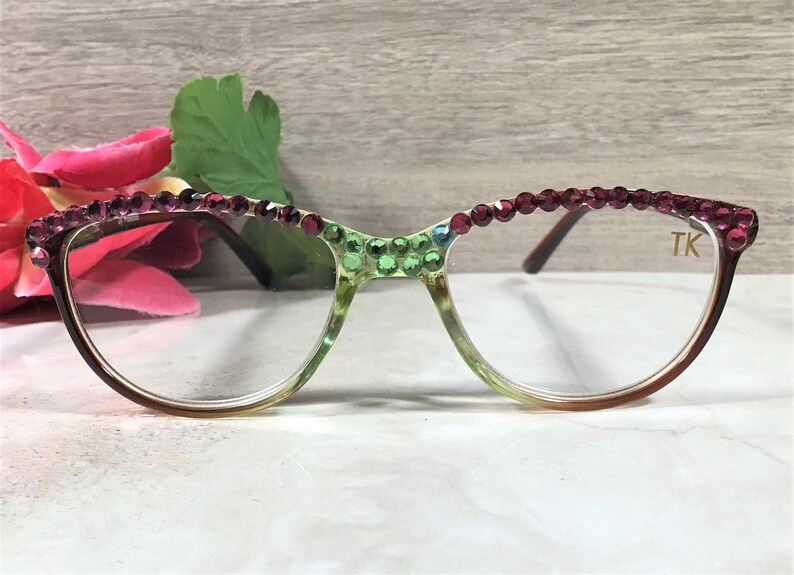 May include: Eyeglasses with a gradient frame, adorned with sparkling rhinestones in shades of green and burgundy. The lenses are clear, and the right temple features the initials "TK". The glasses are set against a soft background with floral accents.