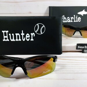 May include: Two pairs of black sunglasses with orange mirrored lenses. The sunglasses are in their cases, which are black with white text and a baseball graphic. The text on the cases reads "Hunter" and "Charlie".